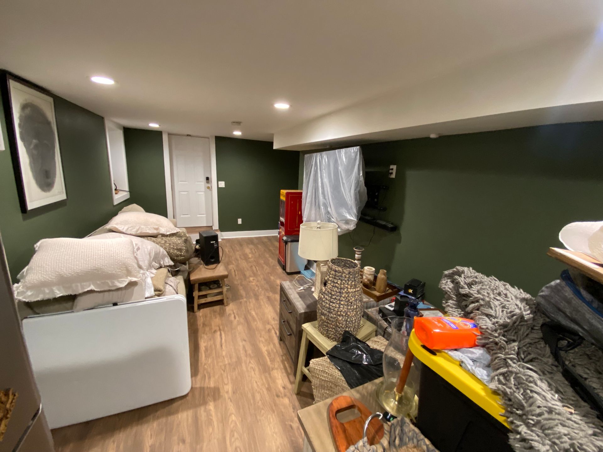 Cluttered basement room with green walls, hardwood-like flooring, and various items, including furniture and storage.