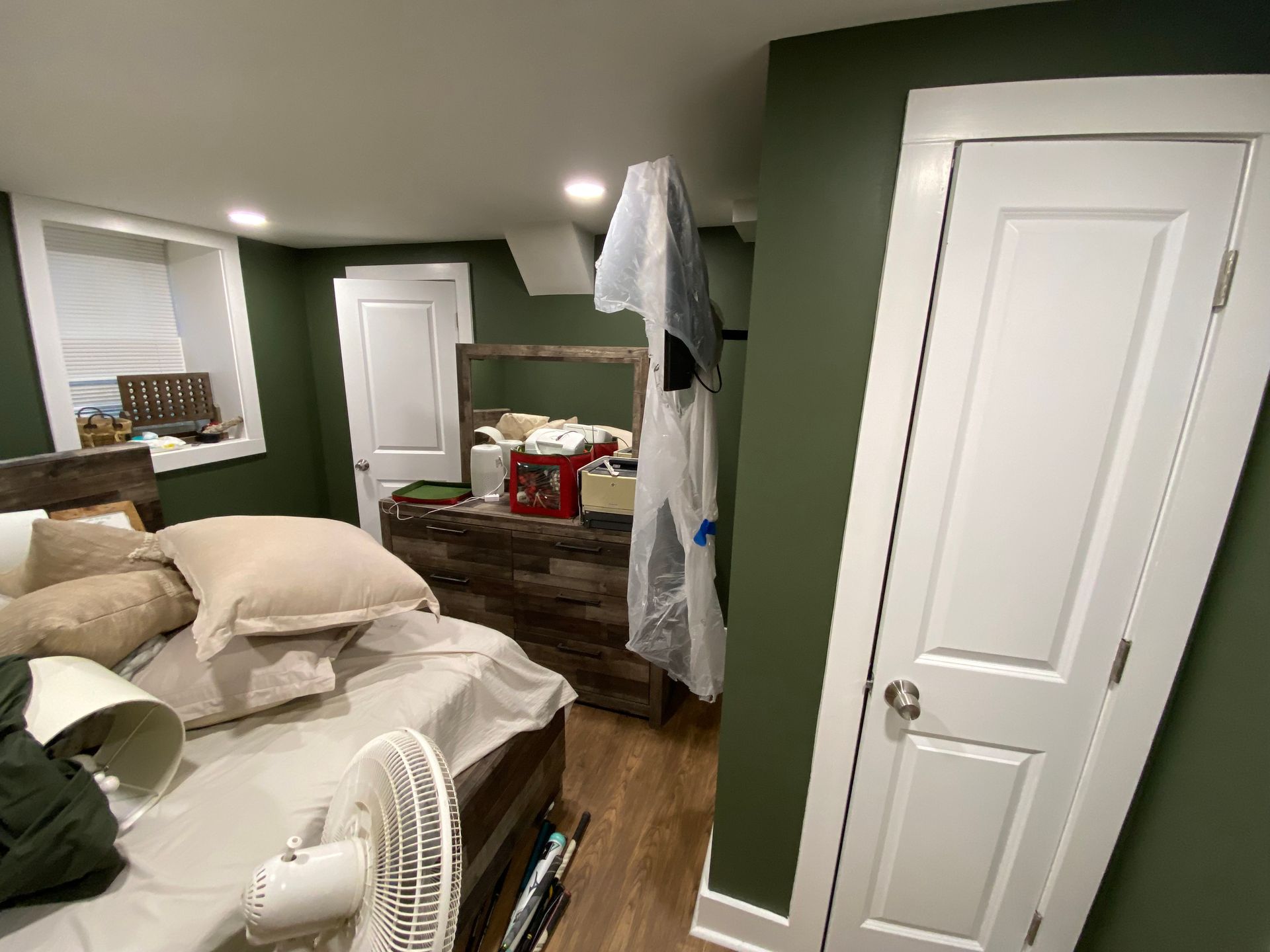 Bedroom with green walls, white door, bed with pillows, dresser, and a window.