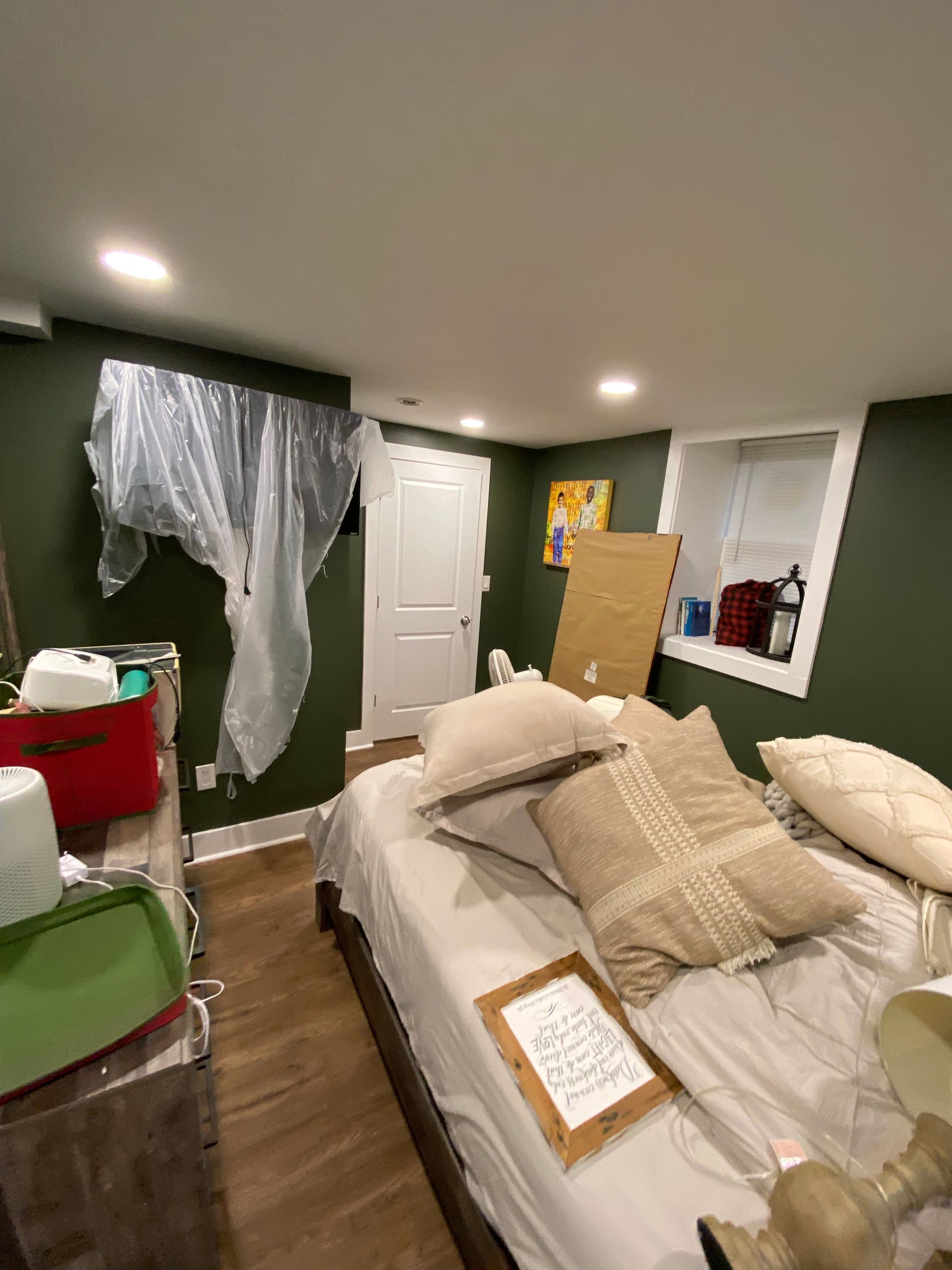 Bedroom with dark green walls, bed with pillows, open door, window, storage containers, and plastic sheeting.