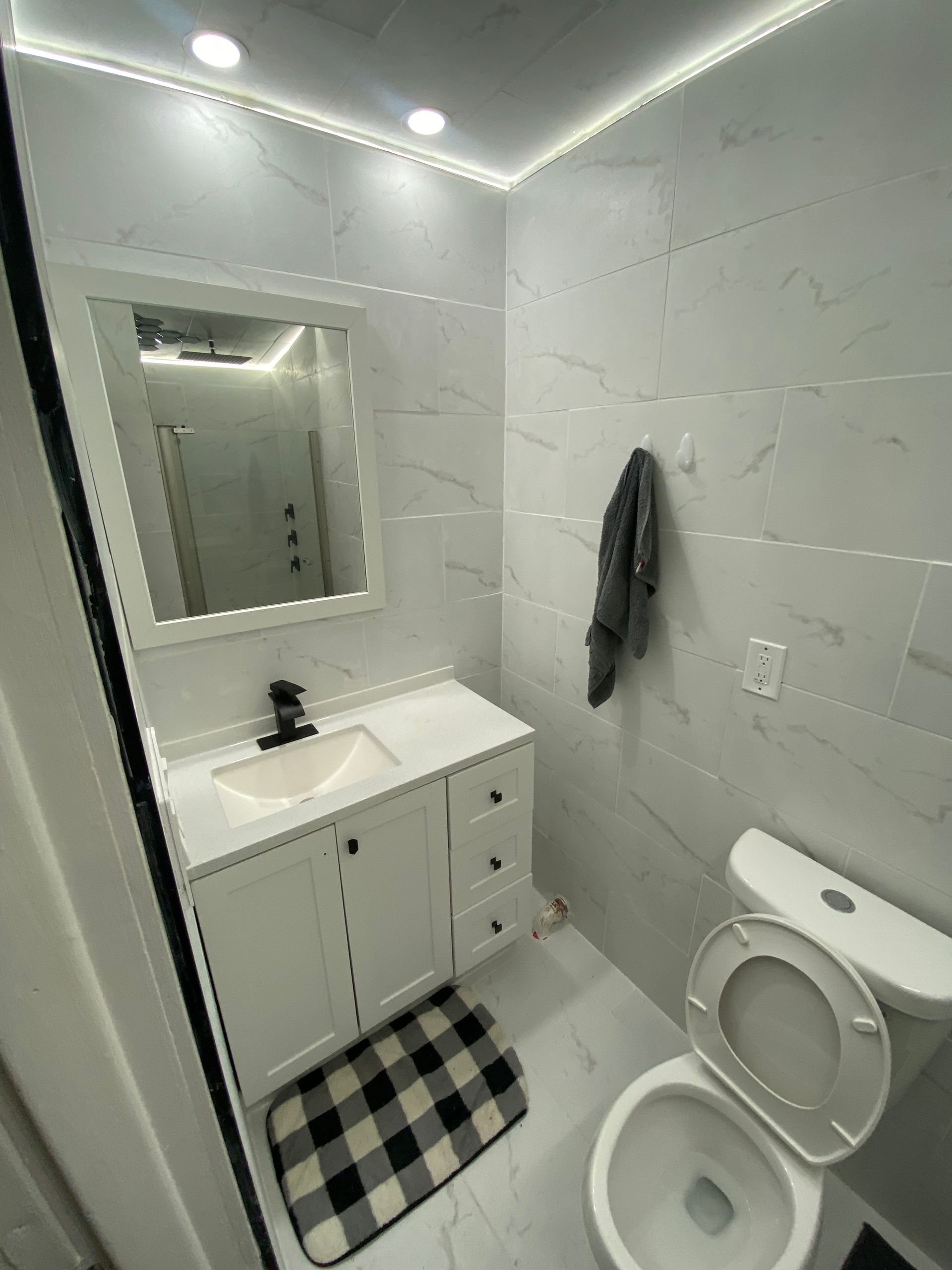 White bathroom with marble-patterned tiles, white vanity, and open toilet seat. Black and white checkered rug.