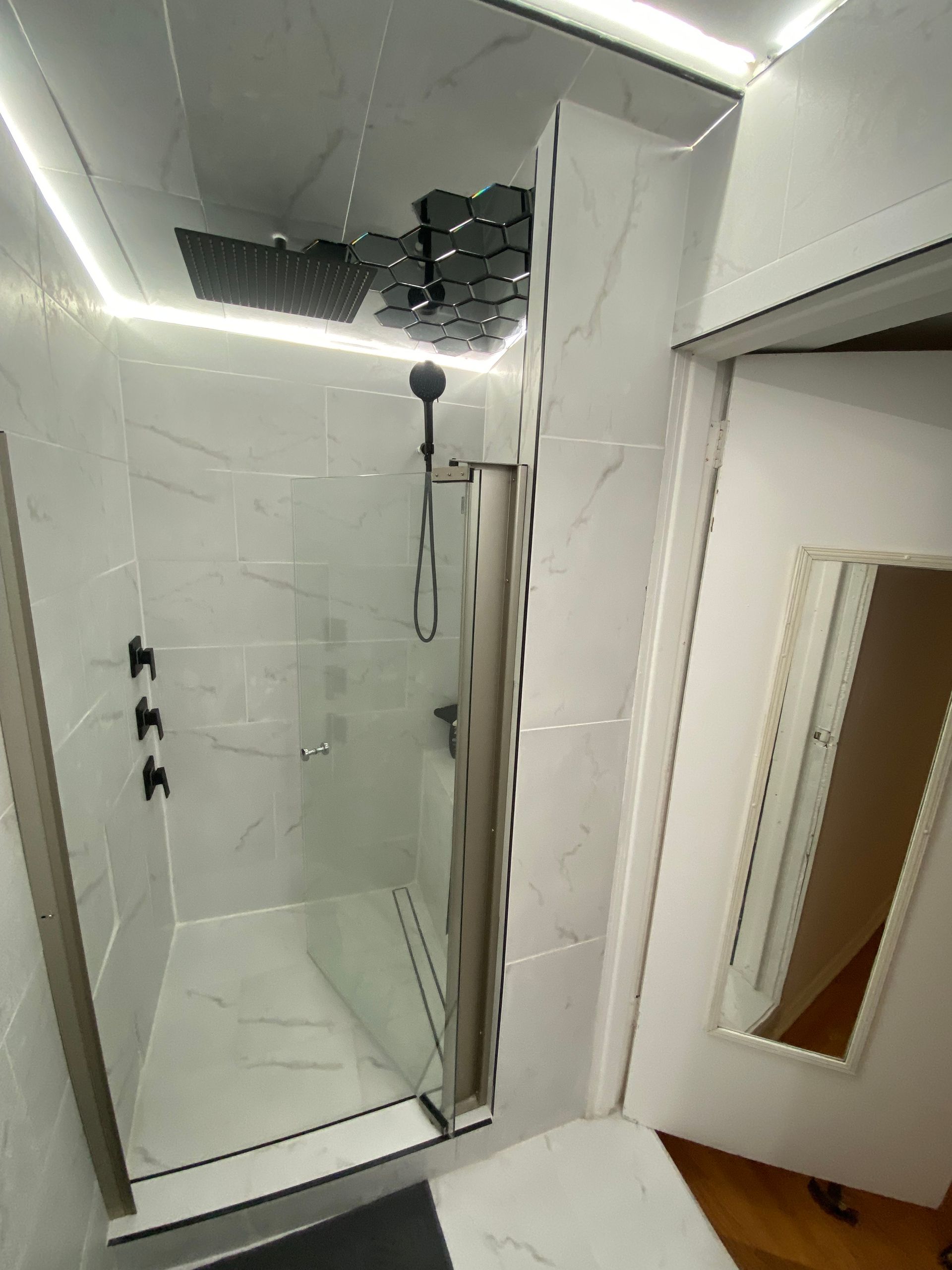 Modern shower with glass door and black fixtures. White marble-look tile, overhead rain shower, and accent lighting.