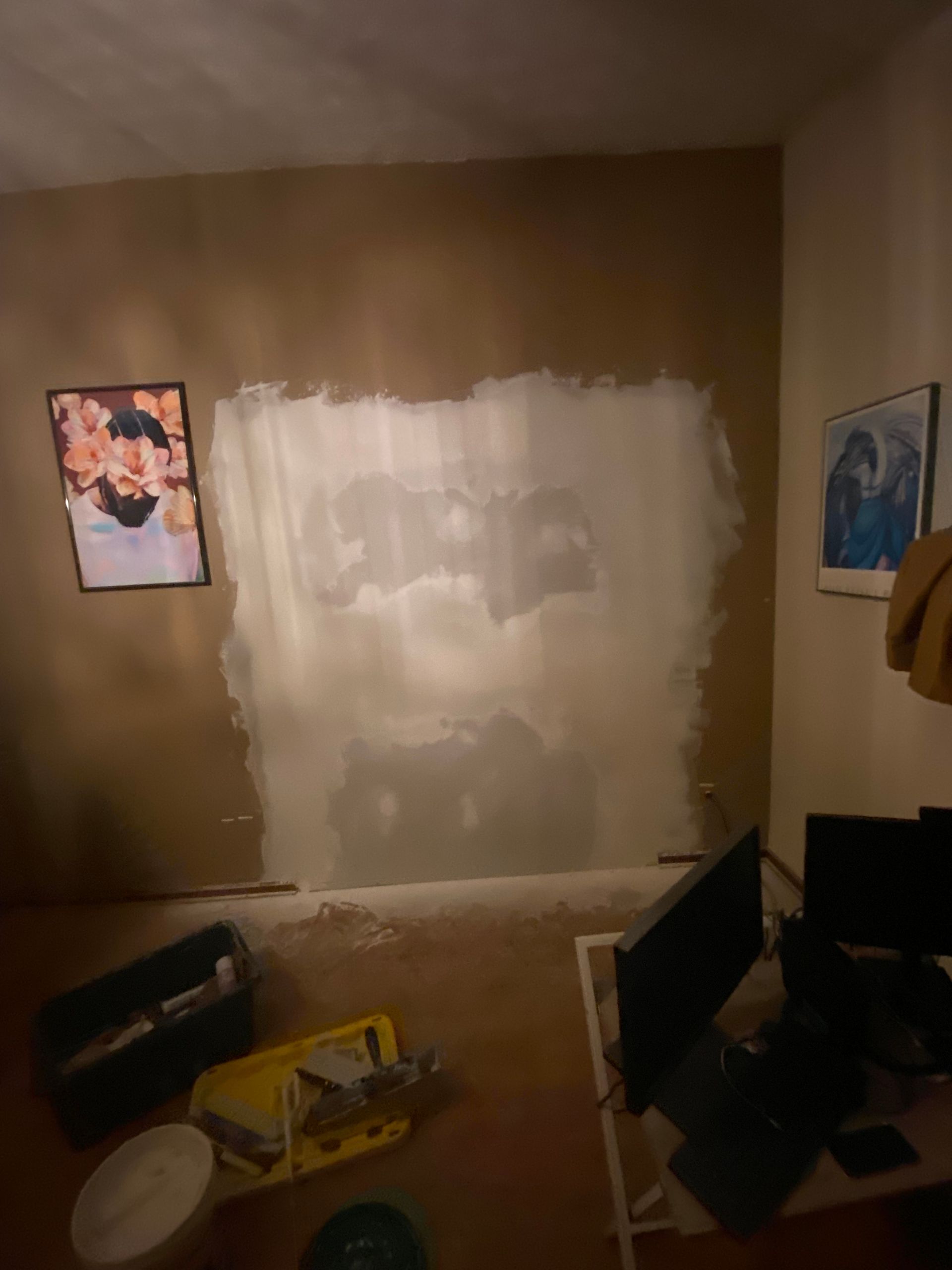 Wall partially painted white, framed pictures, painting supplies, computer monitors on a desk, and debris on the floor.