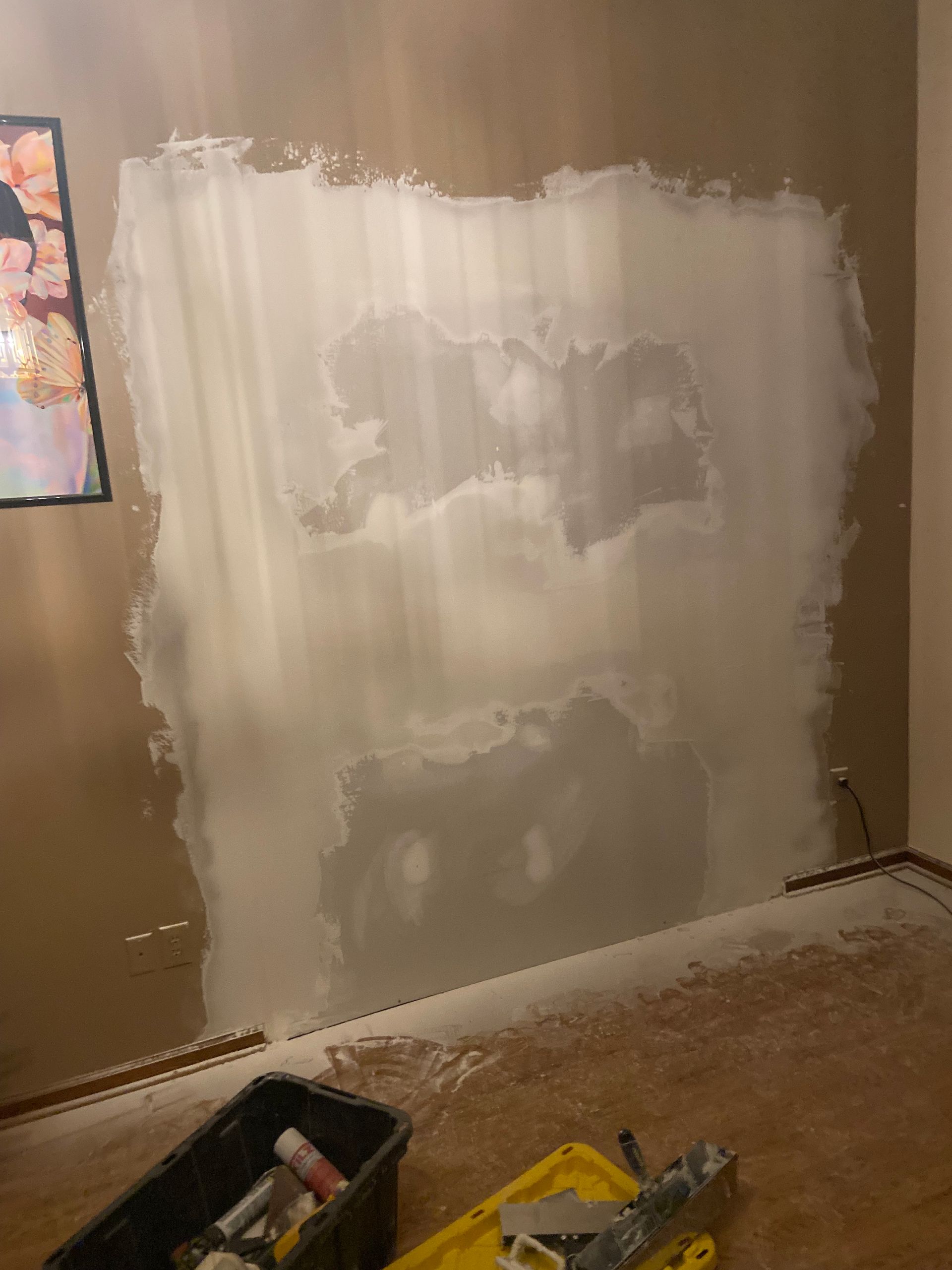 A wall with a large section covered in white compound, tools on floor, next to a painted wall.