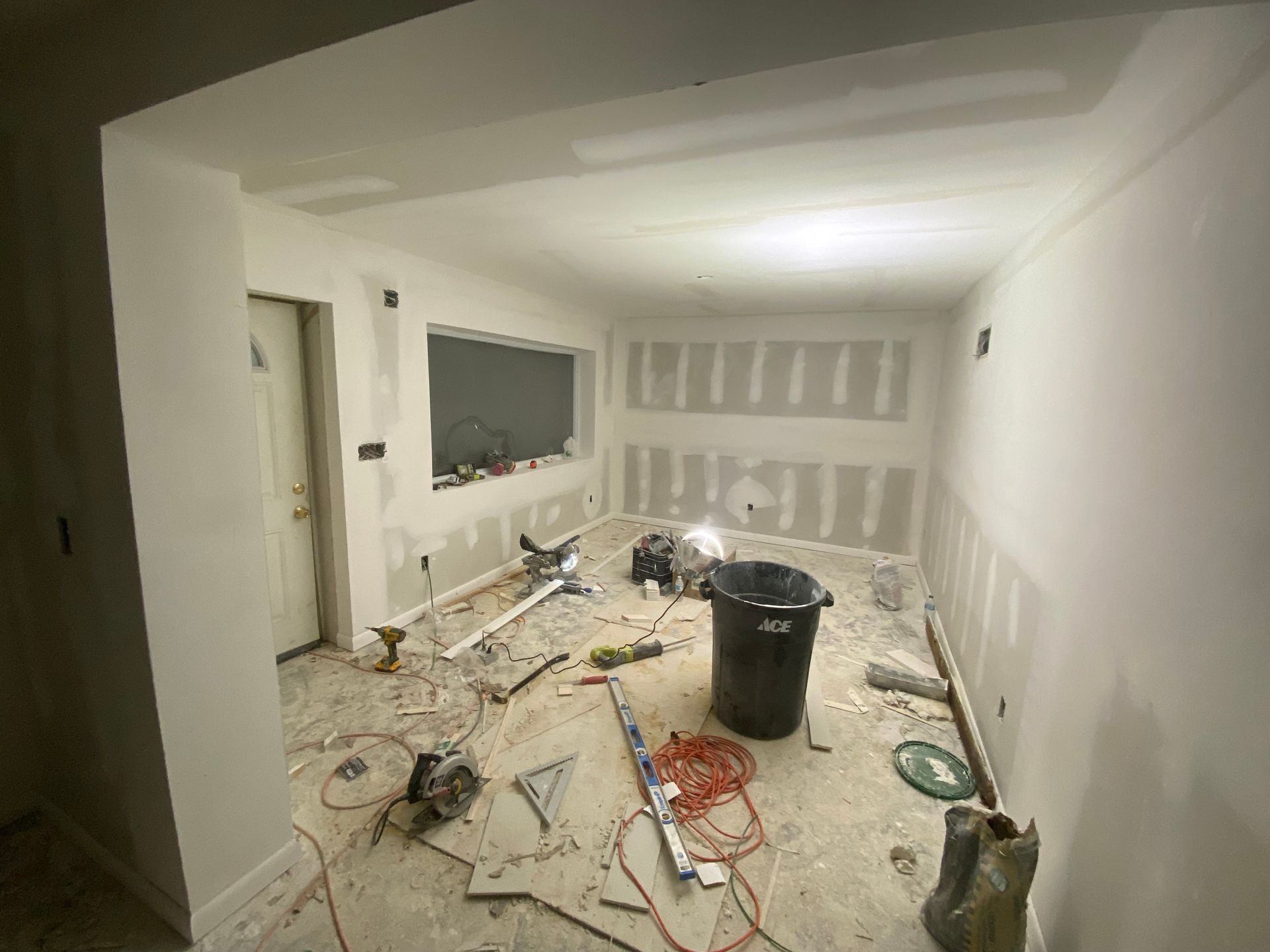 Room under construction with drywall installed on walls and ceiling. Tools and debris scattered on floor.