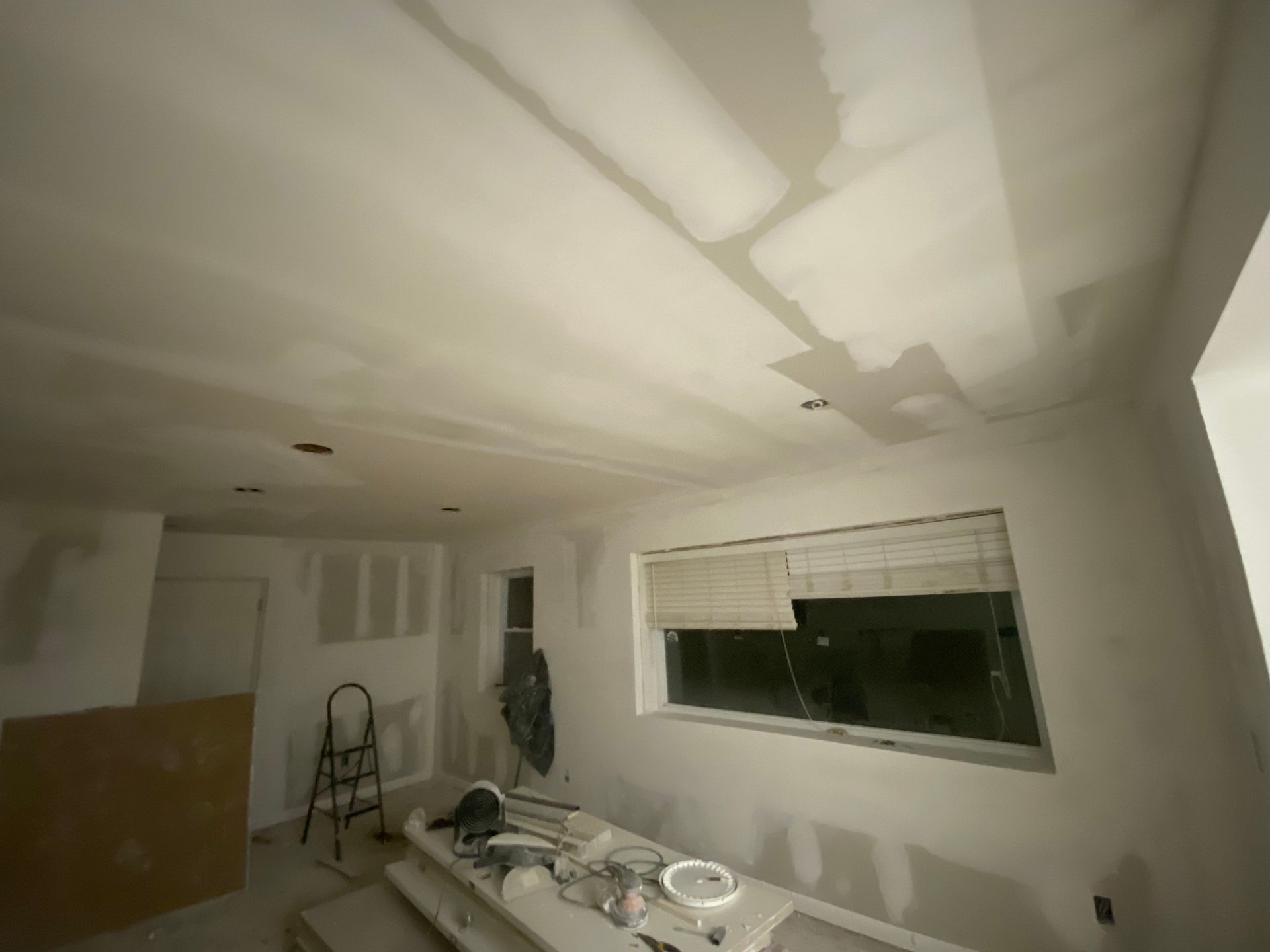 Room under renovation with drywall and unfinished ceiling. Window with blinds, construction tools visible.