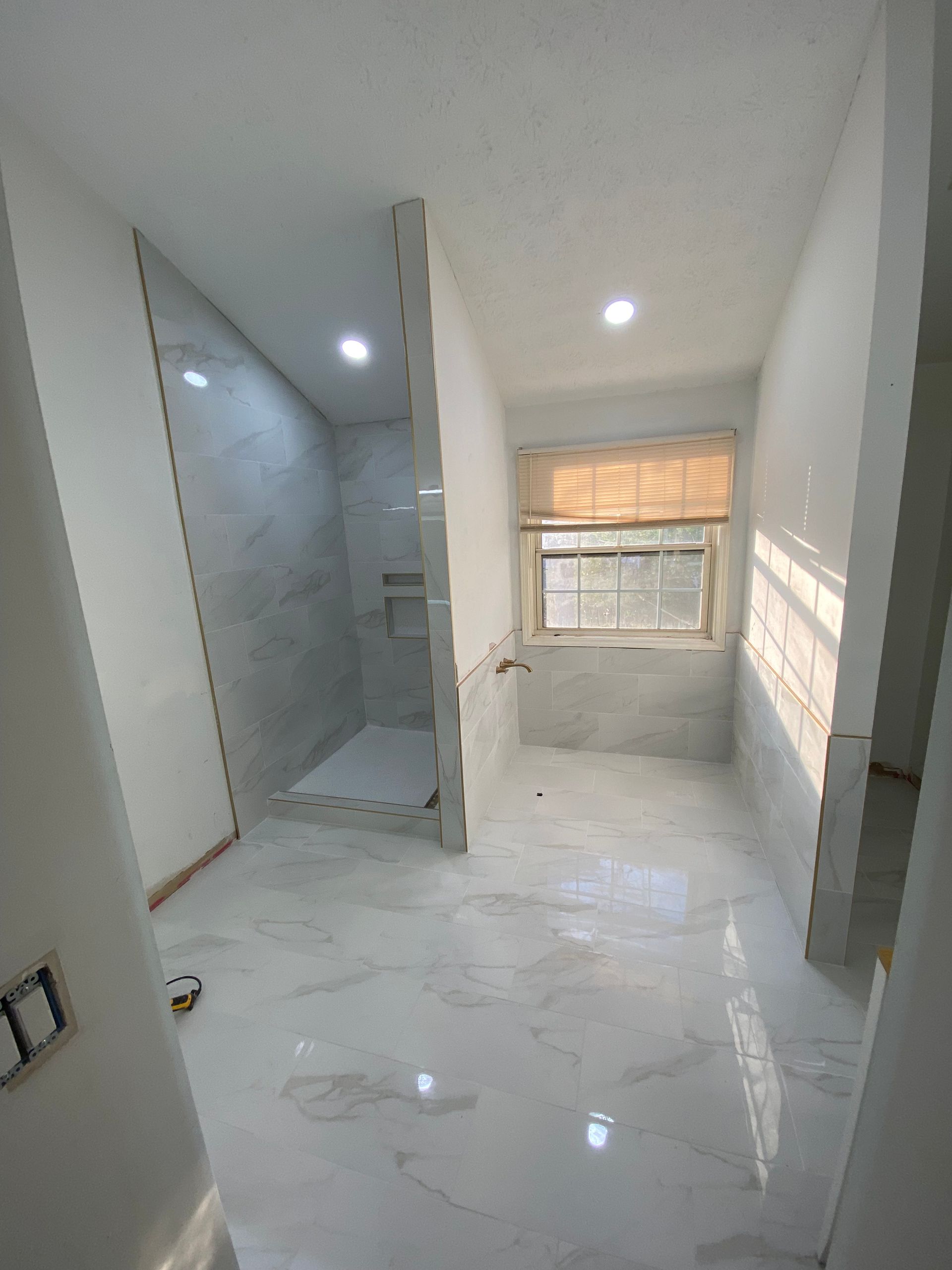 Bathroom under construction, white marble tiles, shower stall, window with blind.