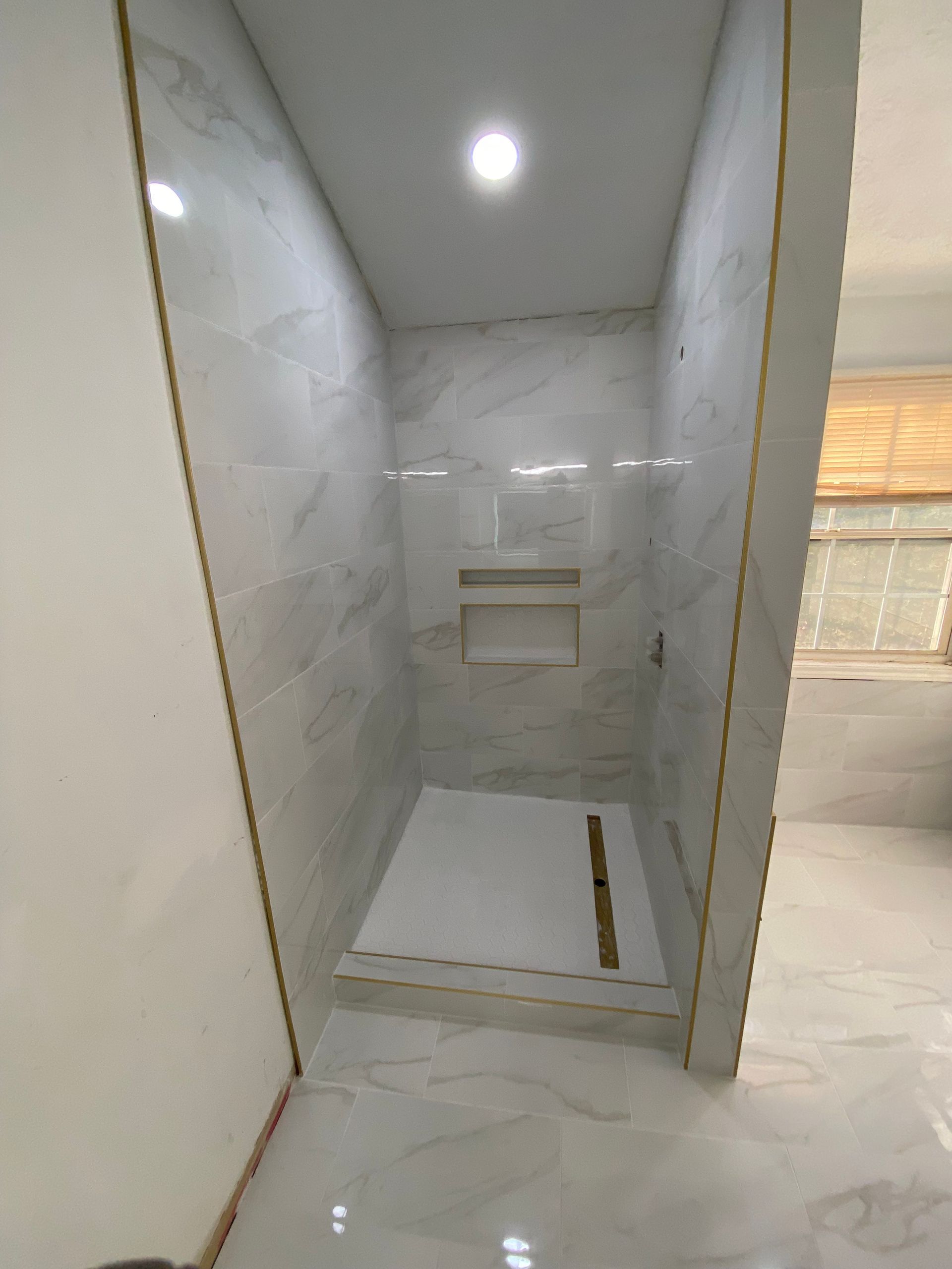 Newly tiled shower stall with white marble-like tiles and recessed shelf.