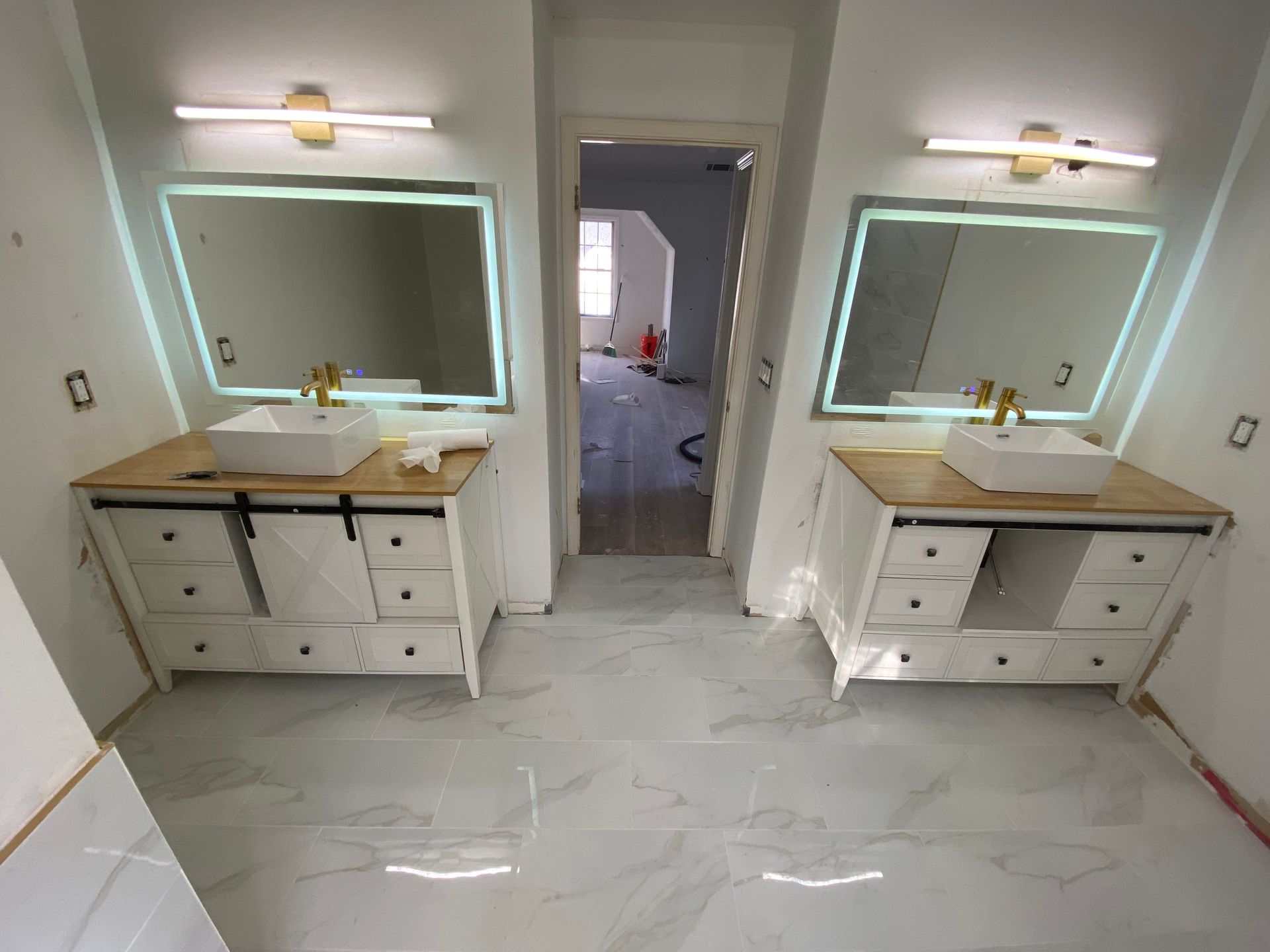 Bathroom with two white vanities, each with a mirror, sink, and gold fixtures. White tile floor.