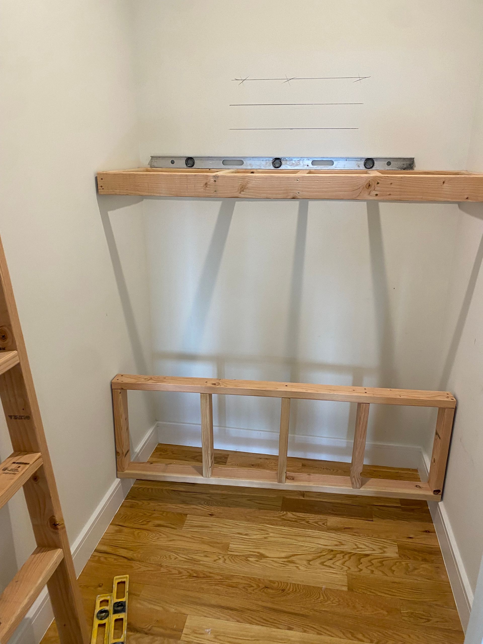 Wooden shelf framework being constructed in a small closet; unfinished wood, level, white walls, wood floor.