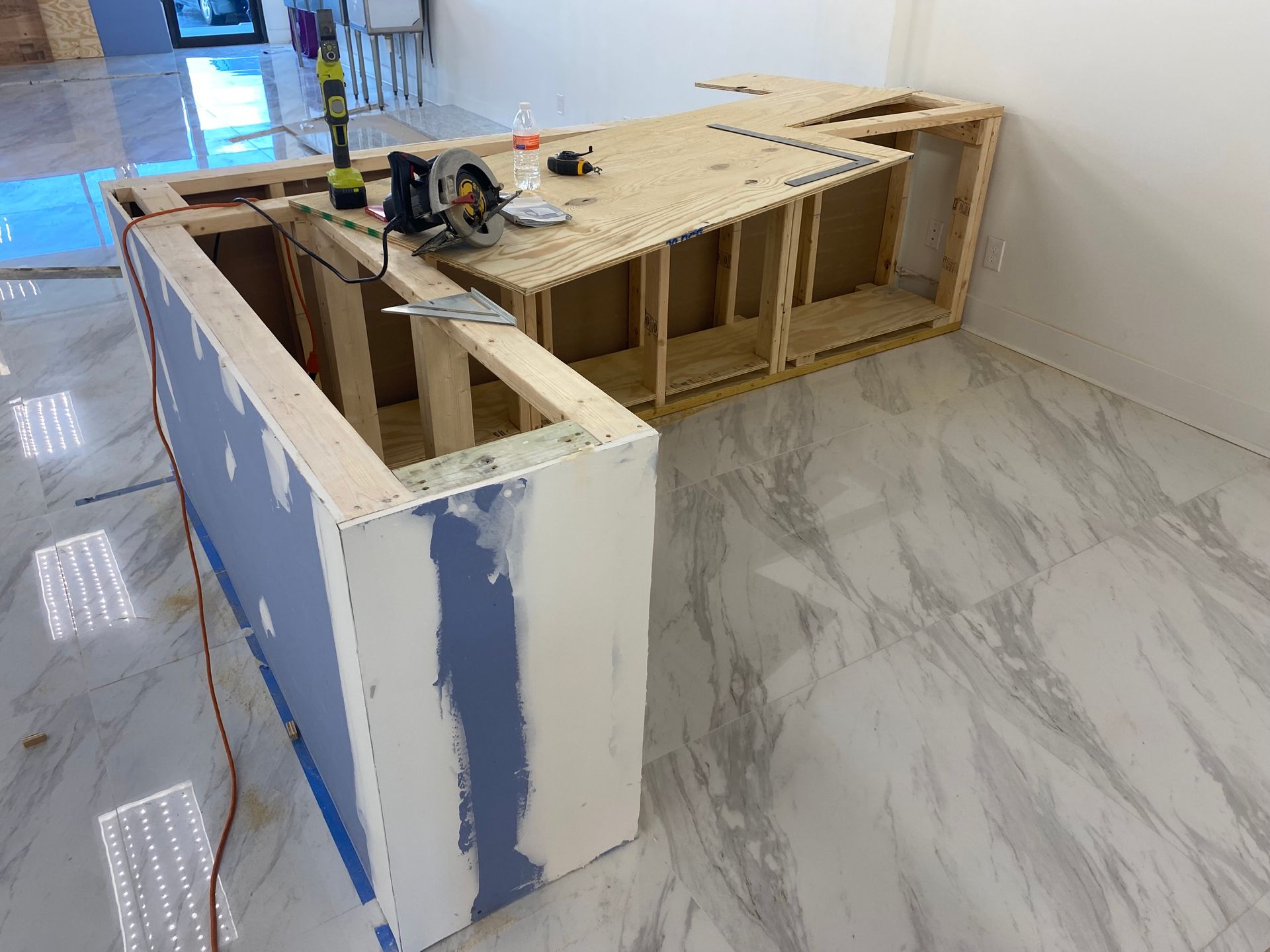 Construction of an L-shaped counter; wood frame, drywall, tools on top, on a tiled floor.