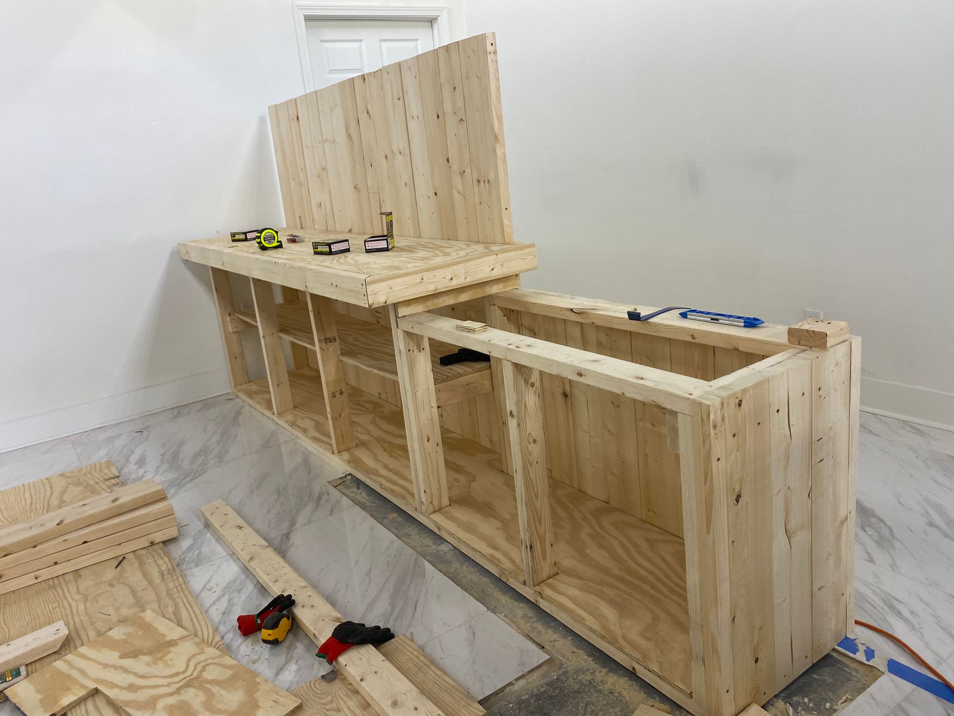 Wooden cabinet under construction in a bright room; various wood pieces, tools, and hardware are scattered around.