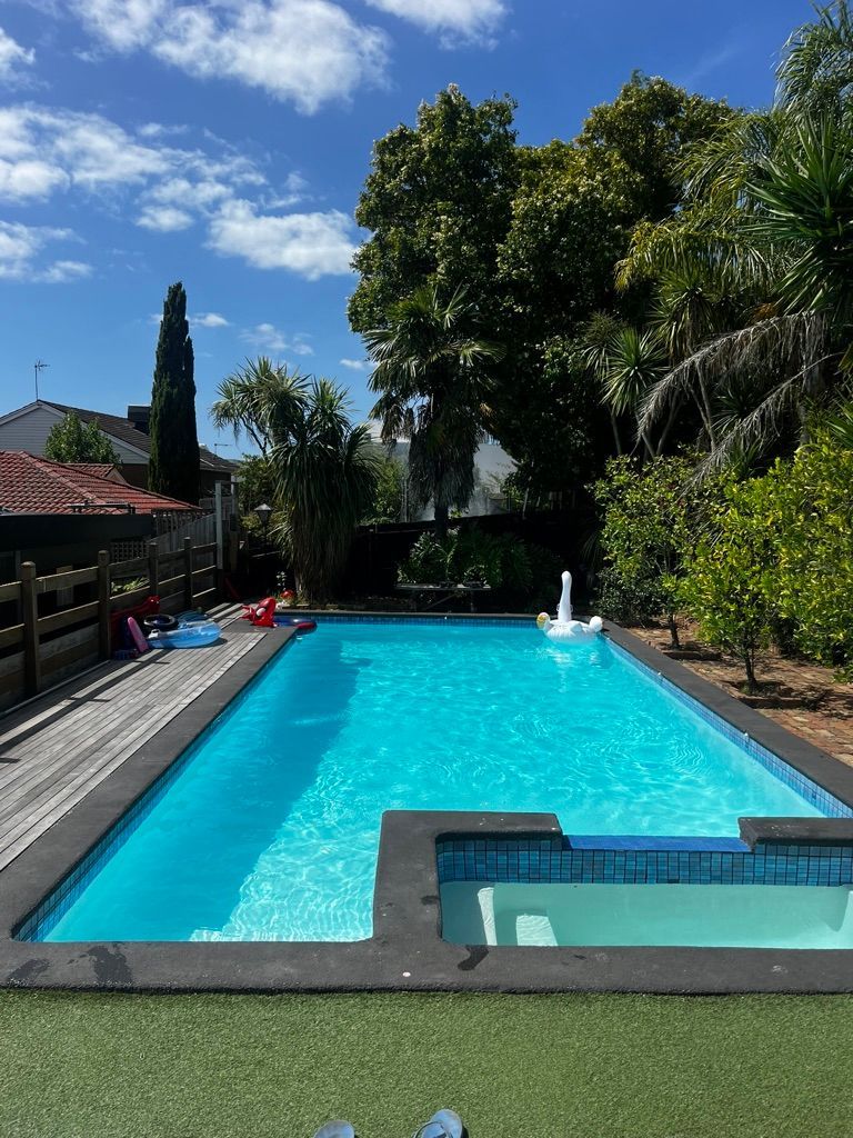 A backyard swimming pool with a smaller attached spa, surrounded by green grass, trees, and a wooden fence under a blue sky.
