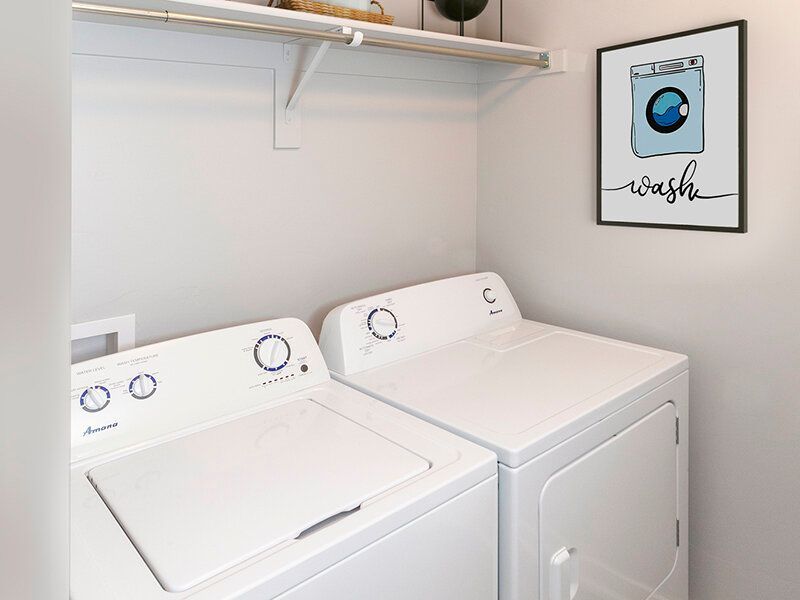 White laundry room with two white top-loading washers and a matching dryer, plus a framed 'wash' art print.