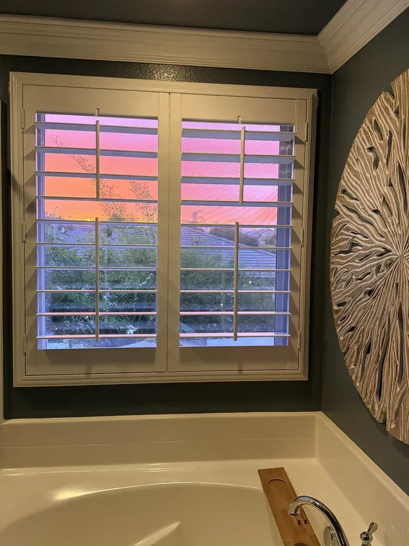 Bathroom window with white shutters, overlooking a colorful sunset. A decorative wall art piece hangs to the right.