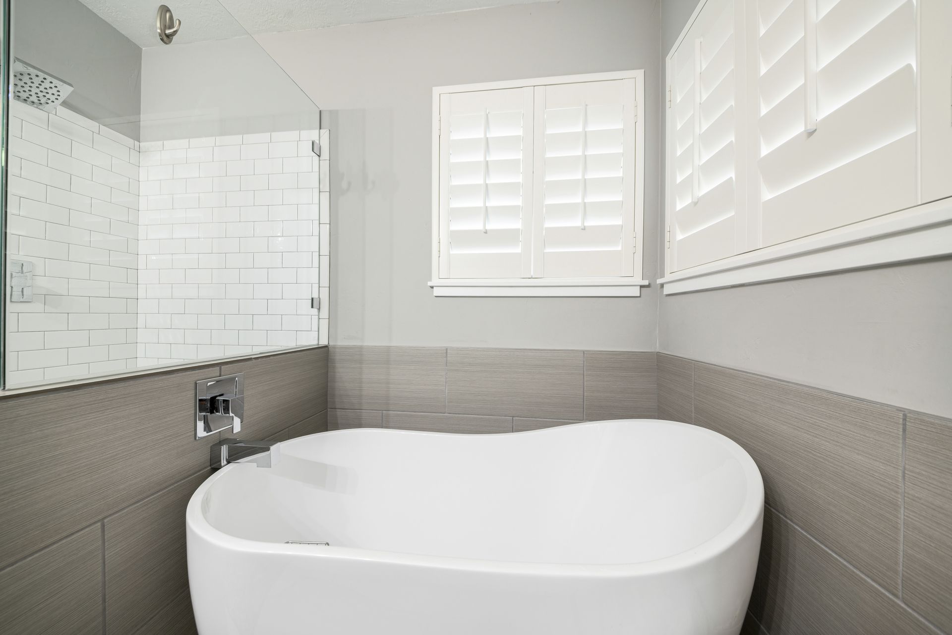 A modern bathroom with a white bathtub, gray tiled walls, and white shutters.