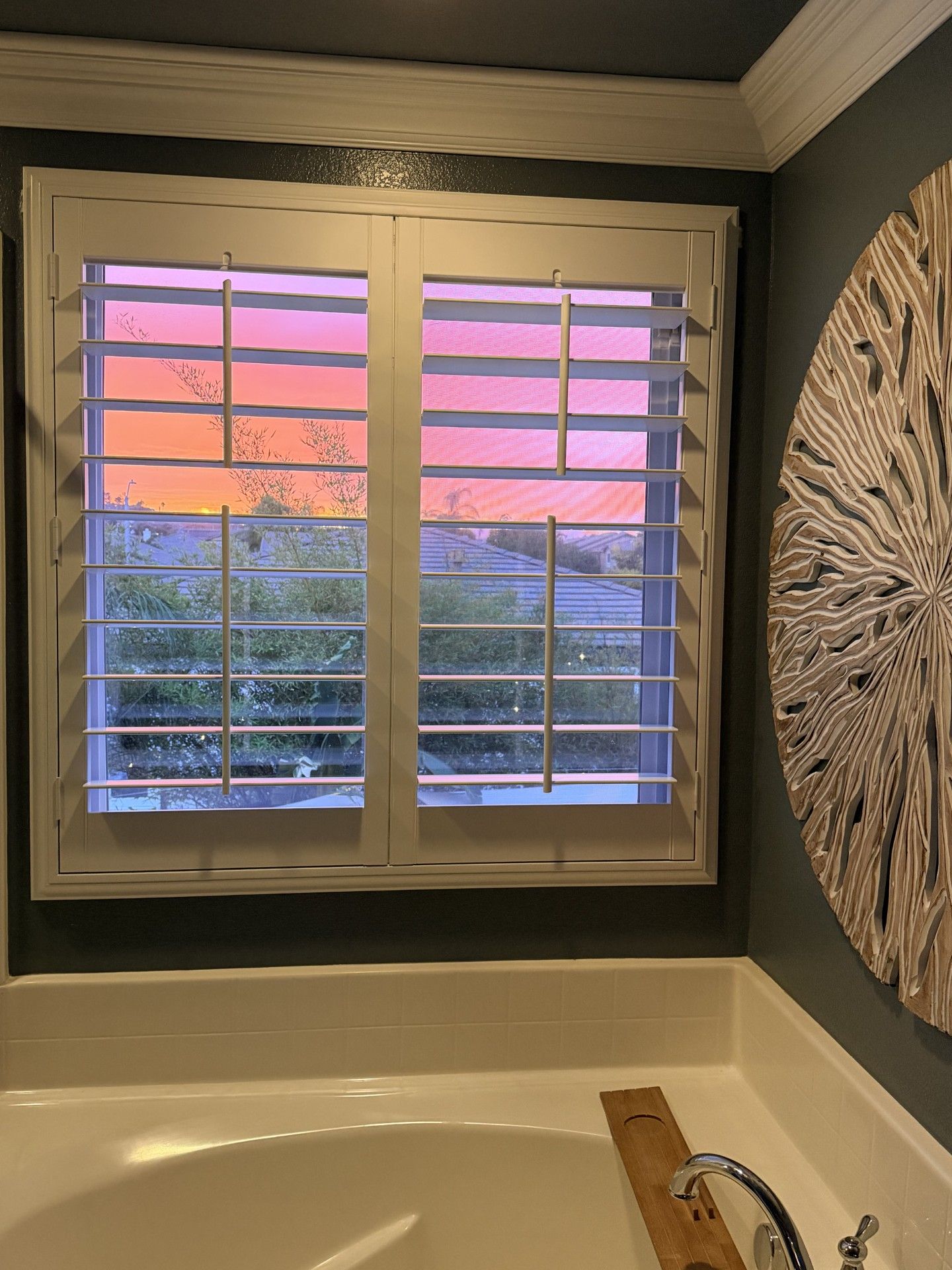 Bathroom window with white shutters, overlooking an orange and pink sunset; decorative wall art to the side.