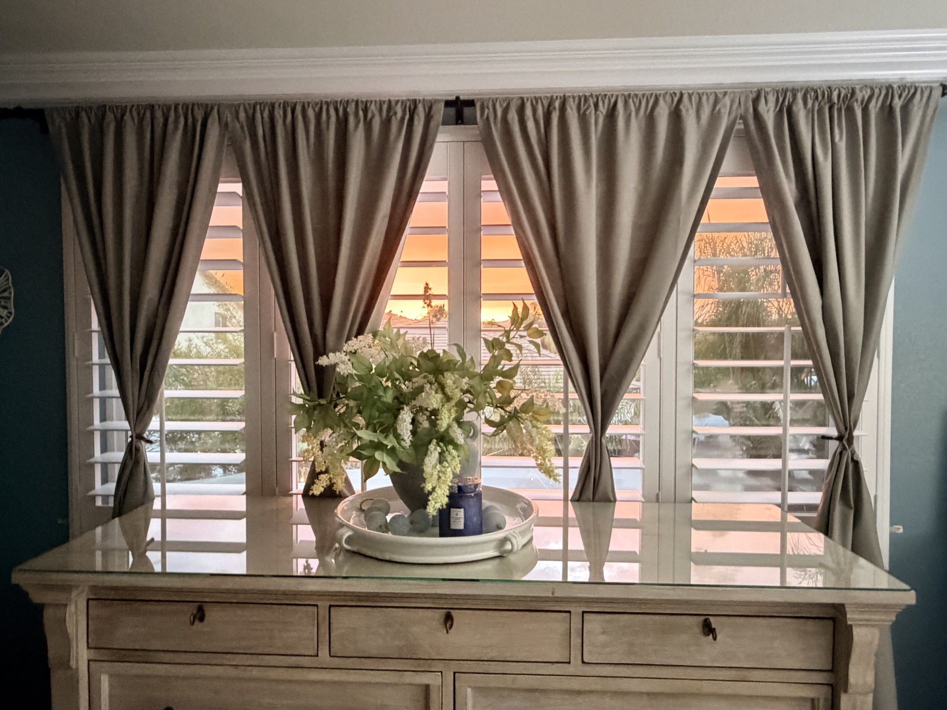 Cream-colored dresser beneath windows with closed white shutters and grey curtains. A flower arrangement sits on top, with a sunset view.