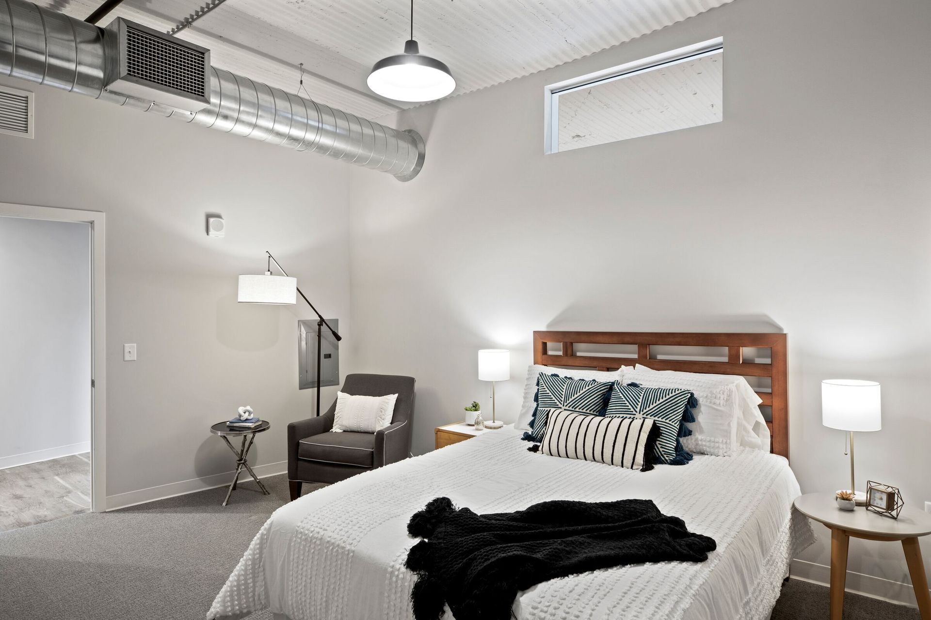 Bedroom in apartment with white bedding, wooden headboard, two nightstands, and exposed metal ductwork overhead.