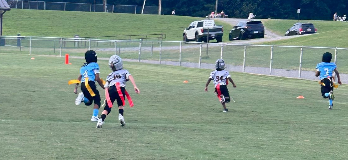 Flag football game at night, a player in red jersey runs with the ball as others pursue on a green field.