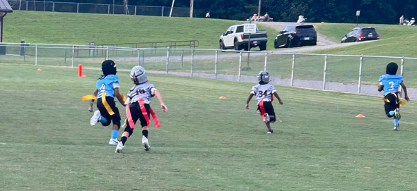 Flag football game at night, a player in red jersey runs with the ball as others pursue on a green field.