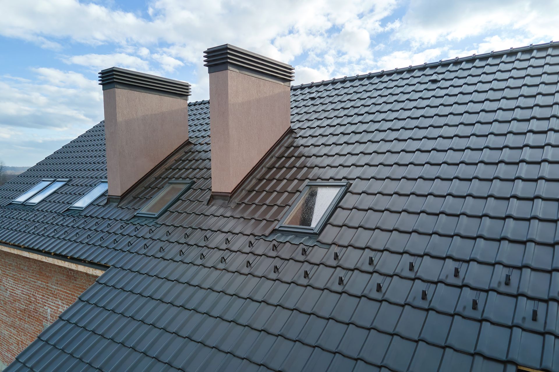 A close-up of a dark tiled roof featuring two beige chimneys and several skylight windows under a cloudy blue sky.