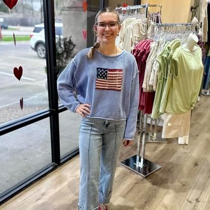 A woman wearing a blue sweater with an american flag on it is standing in a clothing store.