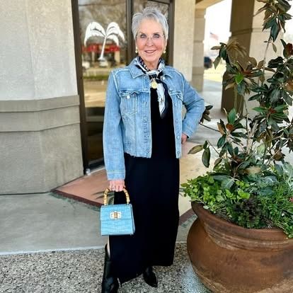 A woman in a black dress and a denim jacket is standing next to a potted plant.