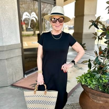 A woman wearing a hat and sunglasses is standing in front of a building holding a bag.
