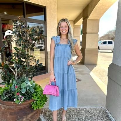 A woman in a blue dress is standing next to a potted plant and holding a pink bag.