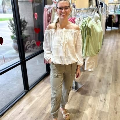 A woman wearing glasses and a white off the shoulder top is standing in a clothing store.