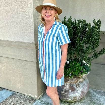 A woman wearing a striped dress and a hat is standing next to a potted plant.