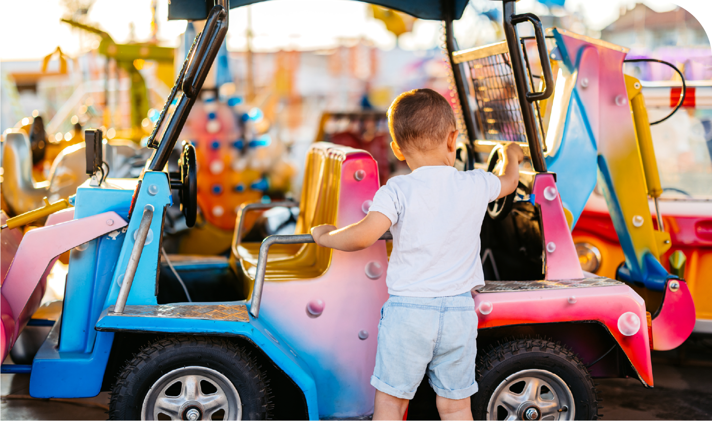 A little boy is standing next to a toy car at an amusement park.