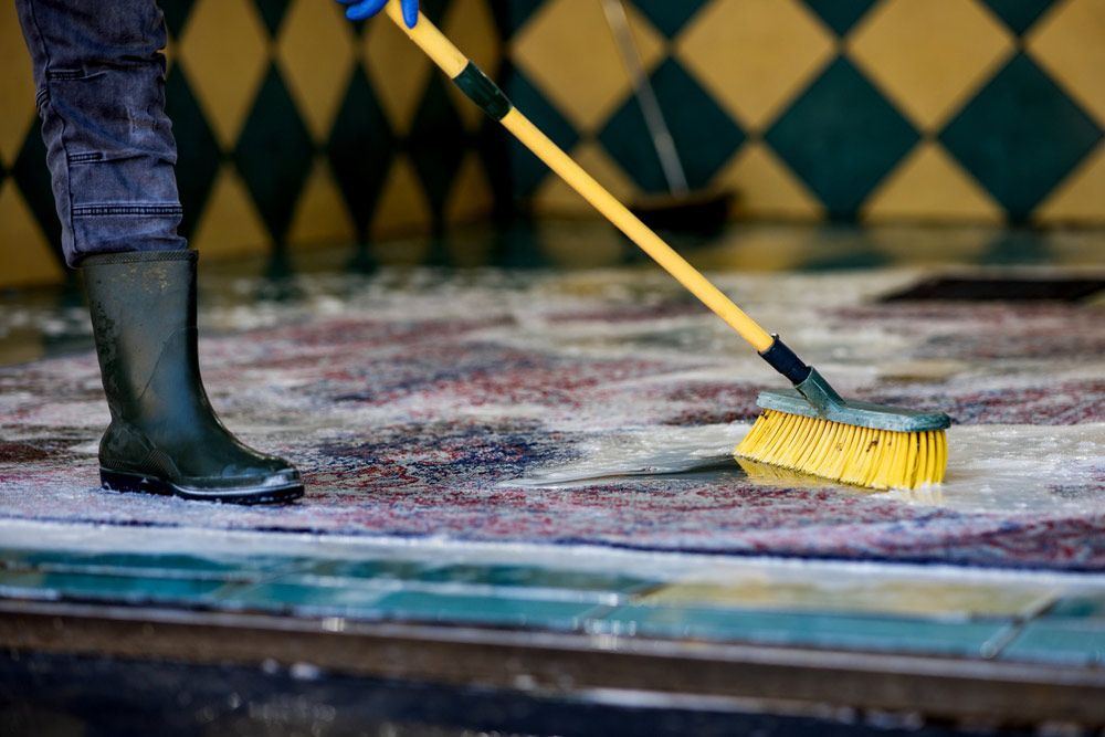 Person in Boots Scrubbing a Wet Carpet With a Yellow Brush in a Checkered-tiled Room — DAH Cleaning Services in Atherton, QLD