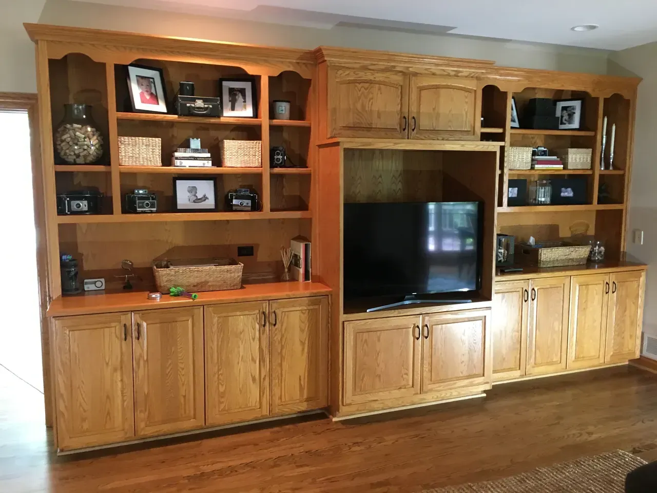 Wooden built-in entertainment center with shelves, cabinets, and a TV. Contains decorative items and rests on wood flooring.