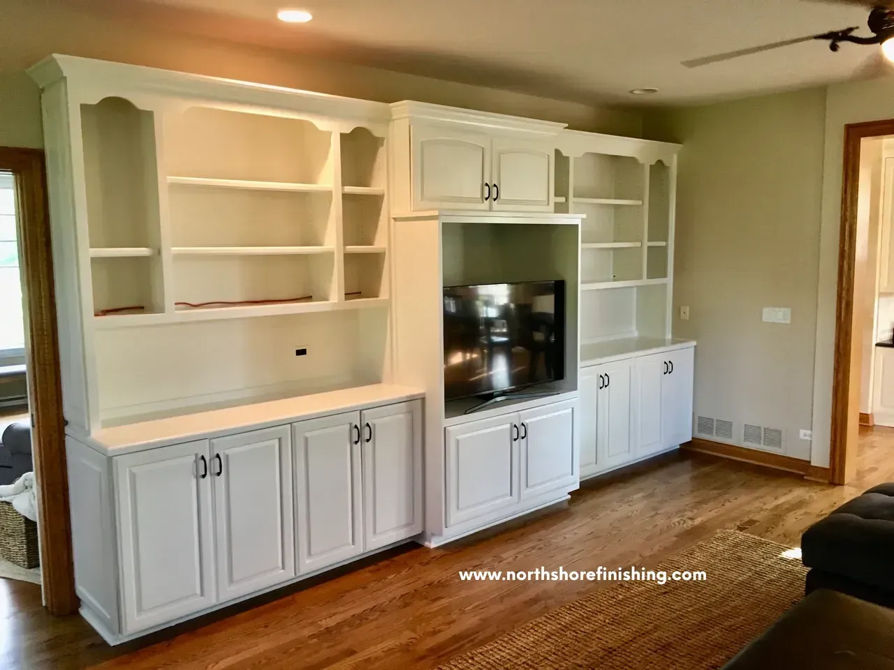 White built-in cabinet with bookshelves, TV, and storage against a light-colored wall and hardwood floor.