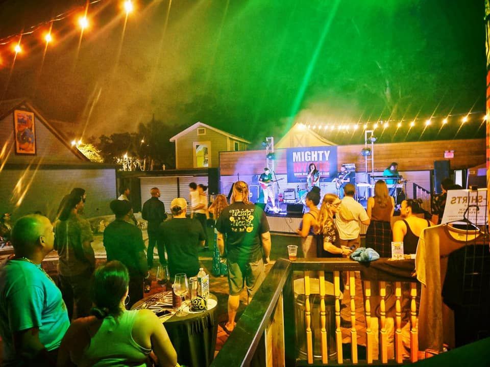 Outdoor concert with band on stage. People watching and dancing under string lights, with green and orange lighting.