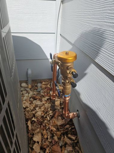 Outdoor utility valve assembly with yellow cap and copper pipes beside a house on gravel.