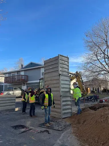 Utility workers in neon vests at a residential construction site beside stacked concrete slabs and an excavator.