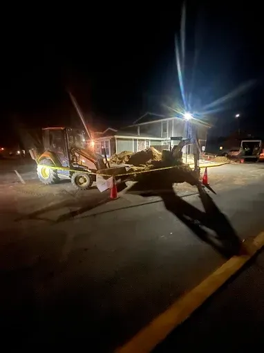 Night roadside construction scene with a backhoe, traffic cones, and bright work lights on a dark street