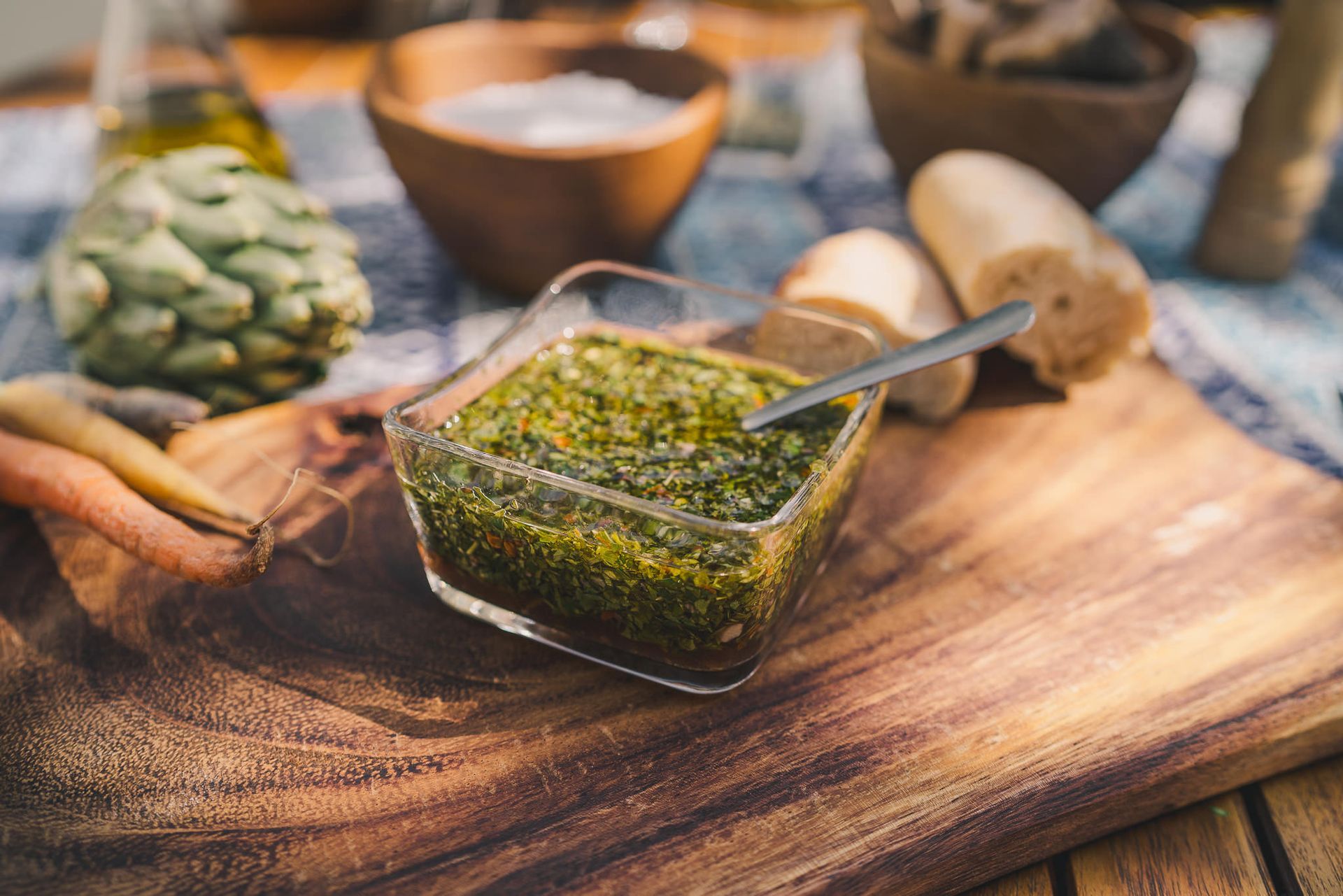 Chimichurri en un plato de cristal con una cuchara sobre una tabla de madera, rodeado de pan y alcachofas.