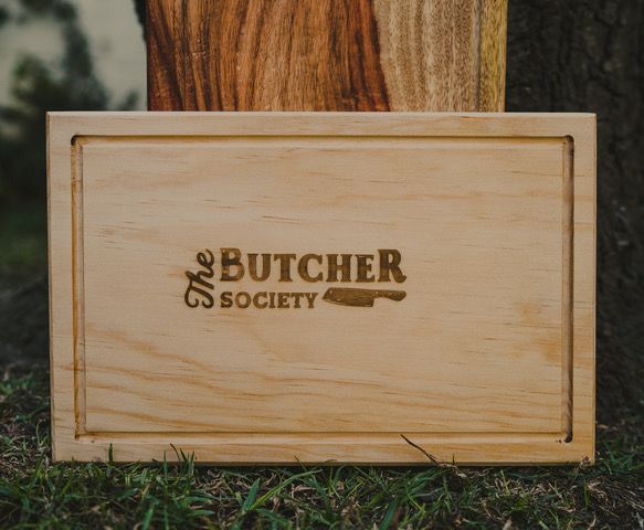 Tabla de picar “The Butcher Society”, apoyada contra un árbol al aire libre sobre el césped.