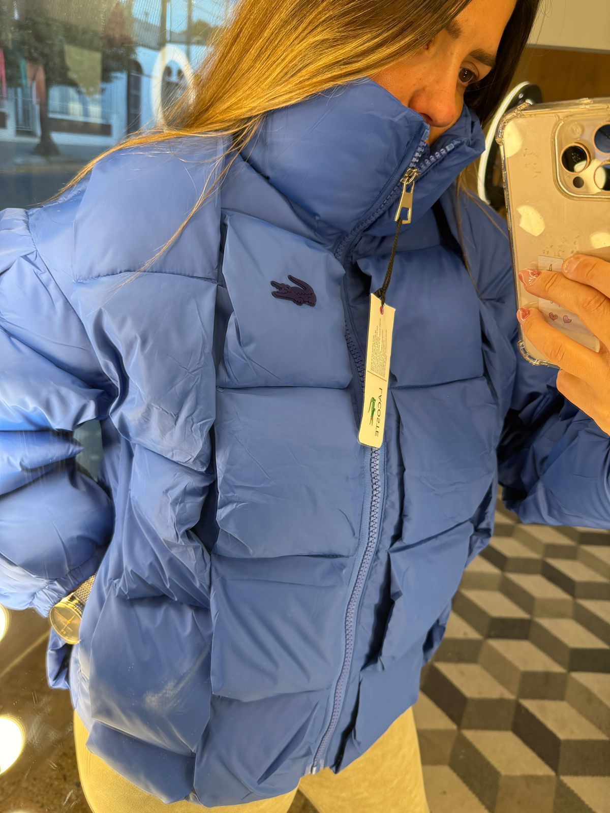 Chaqueta acolchada azul Lacoste con un logotipo azul marino oscuro y una etiqueta con el precio.