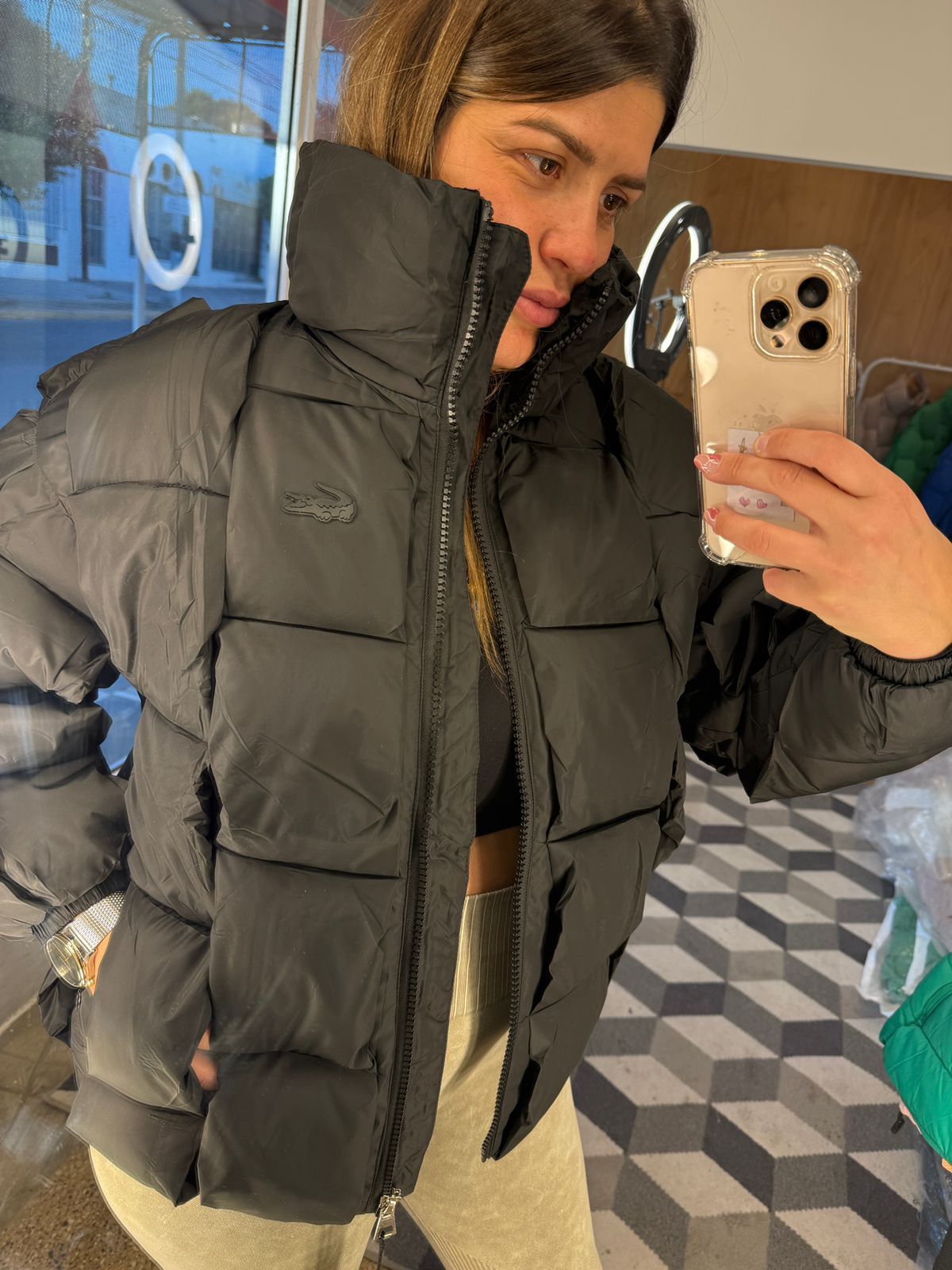 Mujer con una chaqueta acolchada negra de Lacoste, tomándose una selfie, mostrando el logo y la etiqueta con el precio.