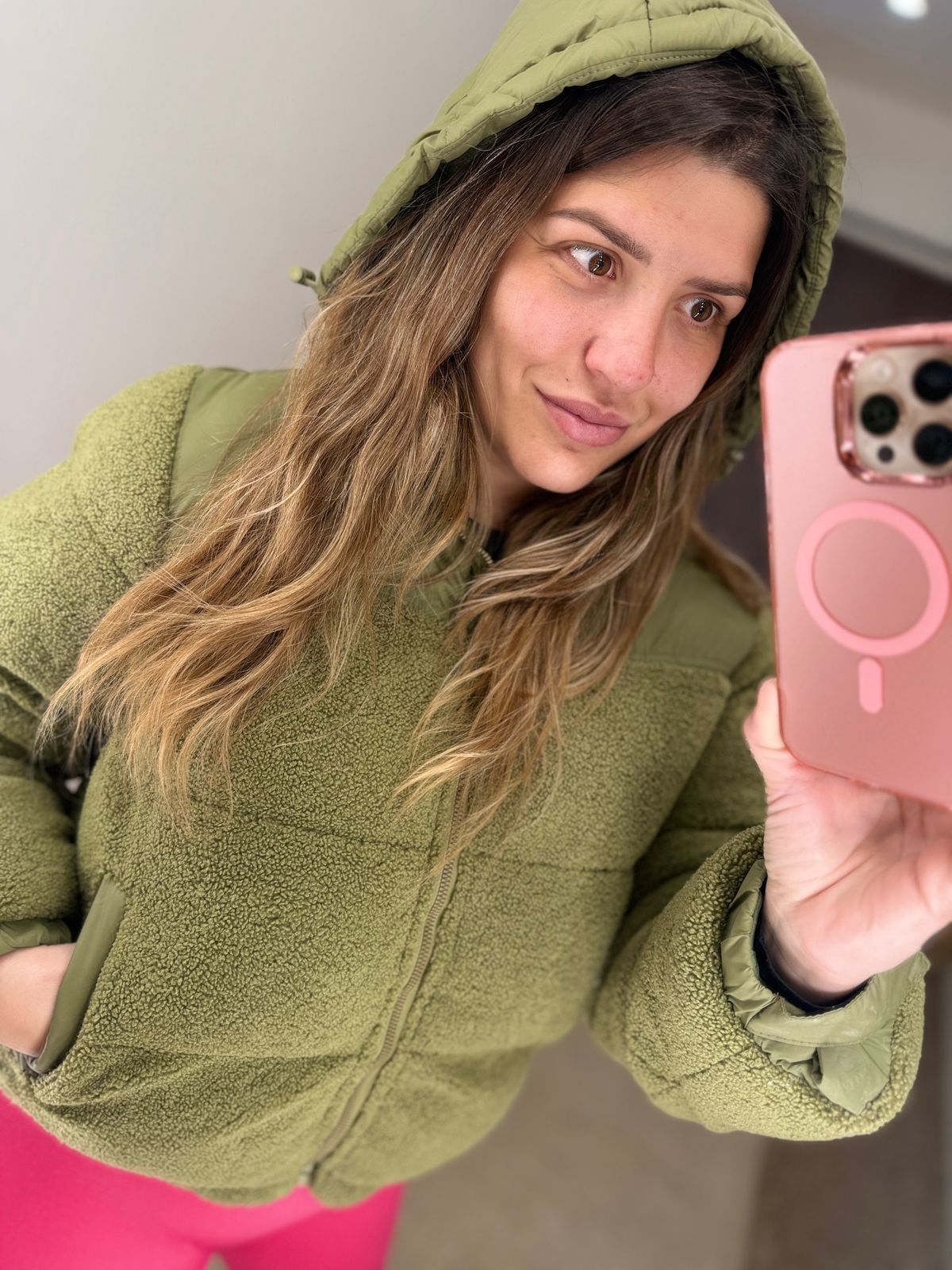 Mujer con una chaqueta verde inflada y leggings rosas tomándose una selfie con una funda de teléfono rosa.