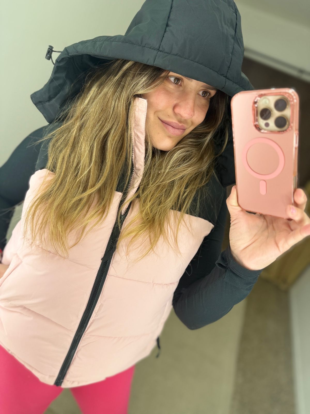 Mujer con chaleco rosa y top negro tomándose una selfie con una funda de teléfono rosa.