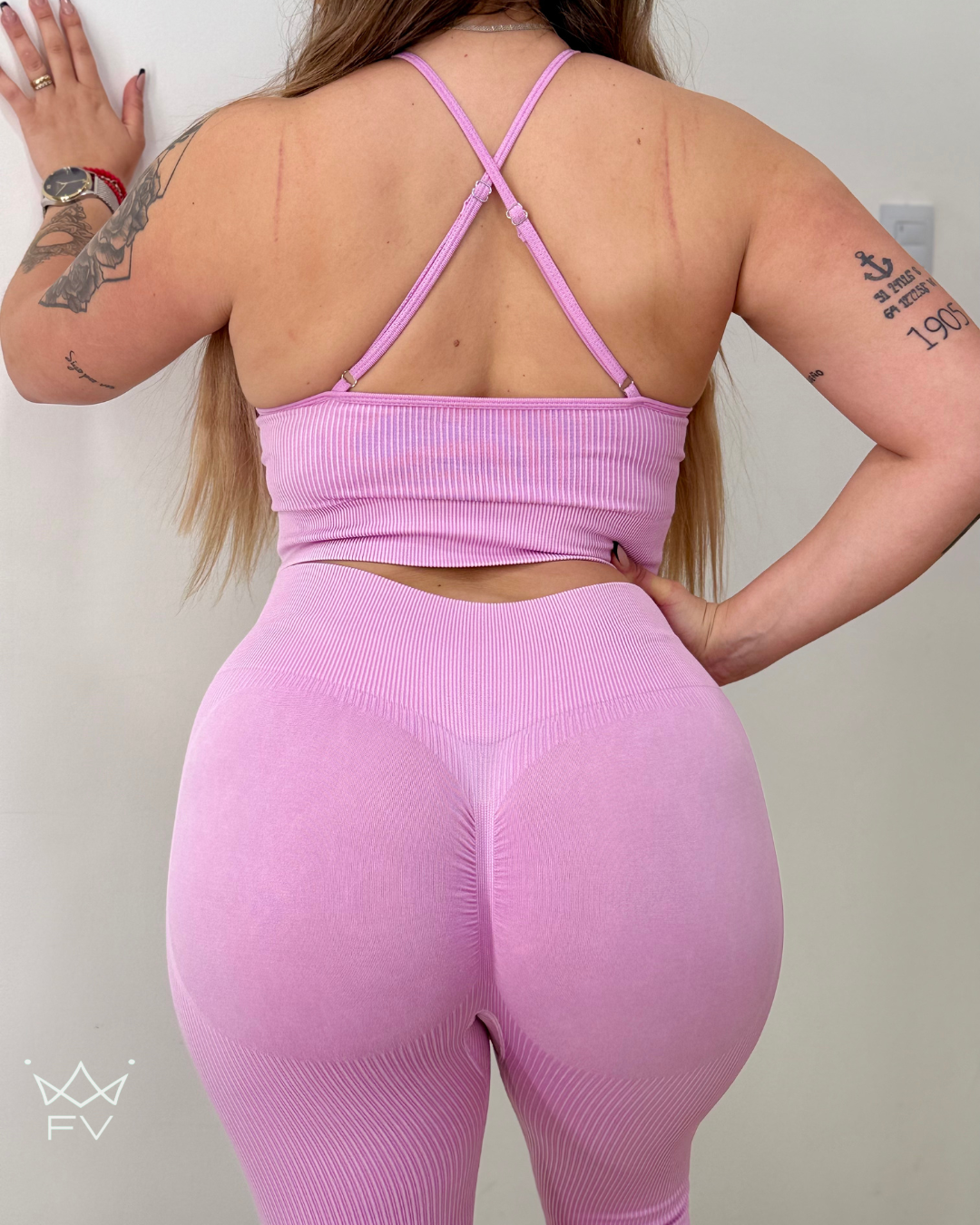 Mujer con un conjunto deportivo rosa, camiseta con cuello en forma de cerradura, de pie frente a una pared blanca.