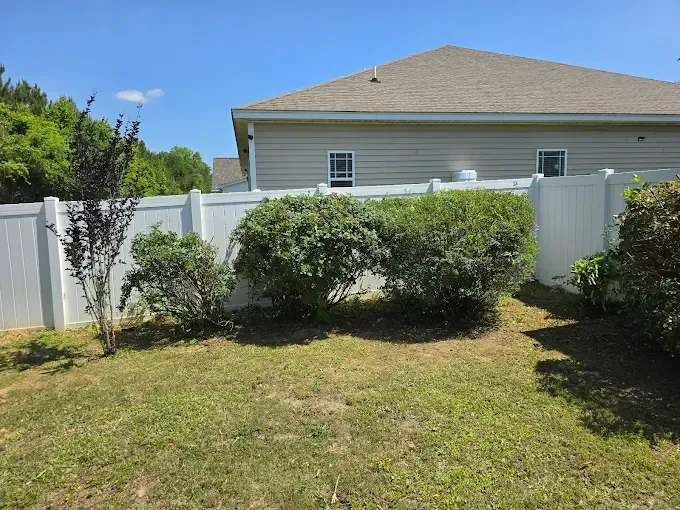White fence and bushes in a yard with a house in the background. Sunny day.