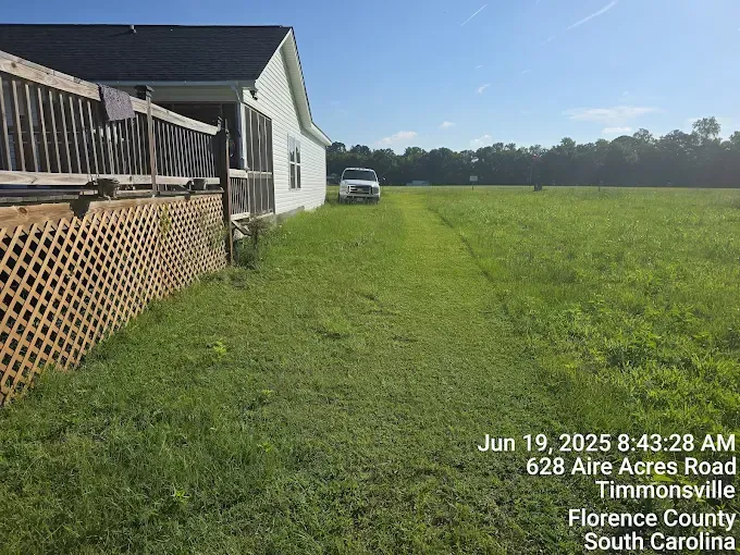 A house with a deck next to a grassy field. A white vehicle is in the field, under a blue sky.