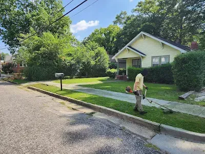 Person trimming grass with a weed whacker on a sunny day in front of a yellow house.
