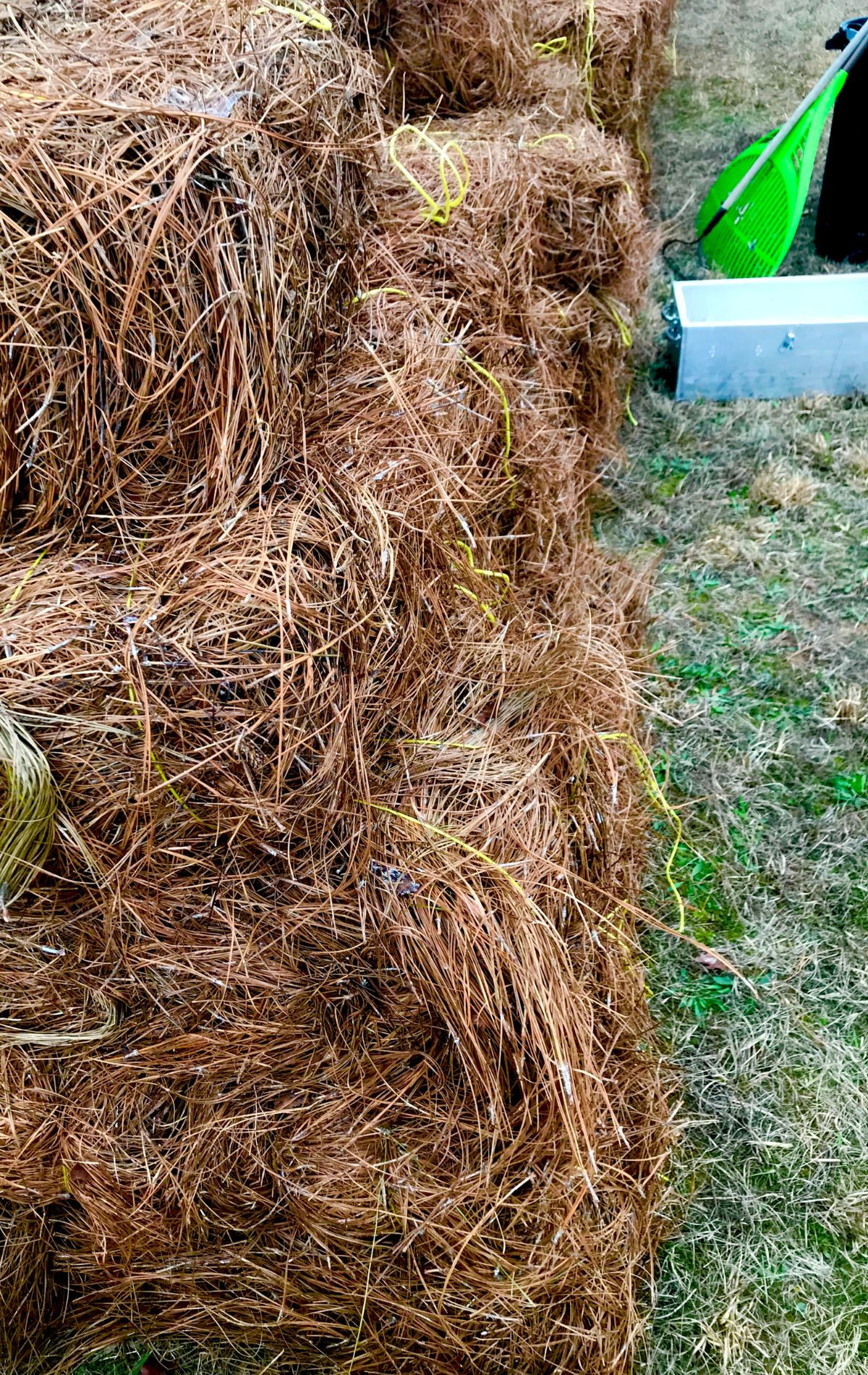 Hay bales stacked in a row on grass, with a green object and concrete block in the background.